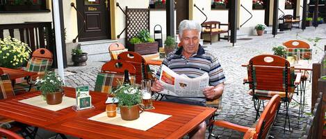 Een man leest een krant op een terras met houten tafels en stoelen, omringd door planten en bloemen in potten.