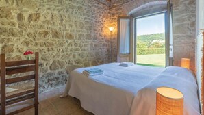 Un dormitorio con paredes de piedra, una silla de madera, una cama con colcha blanca, una mesita de noche y una ventana con vistas a la vegetación.
