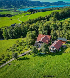 An aerial view of a rural property with two main buildings, a large open field, and a lake in the background. - Bio living Waldblick room with kitchen - Bio Bauernhof Mondsee Familie Schafleitner-Kroiß (3 flowers) (Guggenberg)