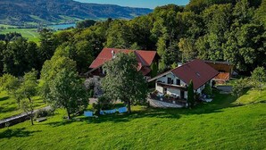 Une maison de campagne avec un toit rouge, entourée d’une végétation luxuriante et avec des collines en arrière-plan.