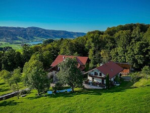 A rural house with a red roof, surrounded by lush greenery and hills in the background. - Bio living Waldblick room with kitchen - Bio Bauernhof Mondsee Familie Schafleitner-Kroiß (3 flowers) (Guggenberg)