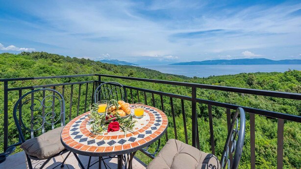 Ein runder Tisch mit Fliesenplatte, umgeben von Metallstühlen, auf einem Balkon mit Blick auf eine üppige grüne Landschaft und ein Gewässer.