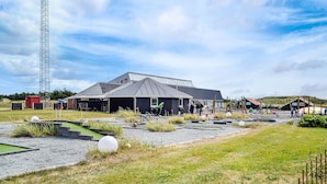 A building with a sloped roof, a fenced area with a small pond, and a tall metal structure in the background. - Lovely apartment in Hvide Sande (Hvide Sande)