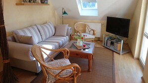 Ein gemütliches Wohnzimmer mit einer Couch, zwei Korbstühlen, einem Couchtisch, einem Fernseher und einem Dachfenster.