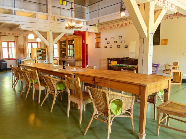 Un grand espace à manger avec une longue table en bois et des chaises en osier, un coin cuisine avec des meubles en bois, et un espace en mezzanine doté d’un plafond en treillis.
