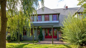 A two-story house with a red door and windows, surrounded by a garden with trees and a wooden pergola. - 270m² apartment - apartment (Rubenow)