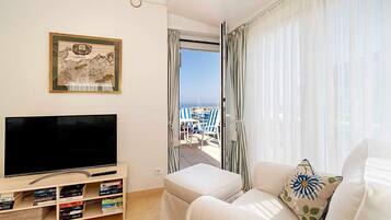 A modern living room with a flat-screen TV, a bookshelf, a sofa, and a balcony with a view of the sea.