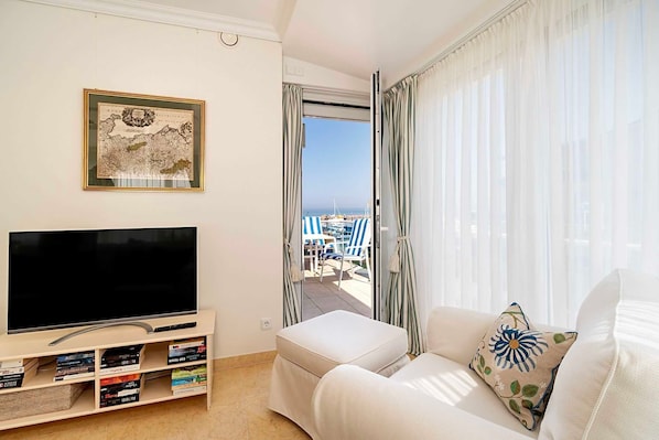 A modern living room with a flat-screen TV, a bookshelf, a sofa, and a balcony with a view of the sea.