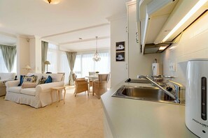 A modern living room with a sofa, dining table, and chairs, a kitchen with a sink and a coffee machine, and a chandelier. - Ut mine Festungstid (Whg. 6) - Strandresidenz-Kühlungsborn ***** (Kühlungsborn)