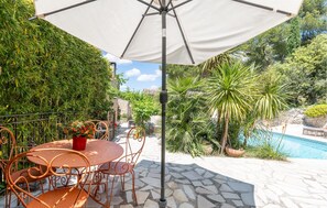 Een buitenterras met een ronde tafel en stoelen, een witte parasol en een zwembad op de achtergrond.