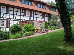Une maison traditionnelle à colombages, avec un mur en pierre, un parasol rouge et un jardin agrémenté de fleurs et d’arbustes.