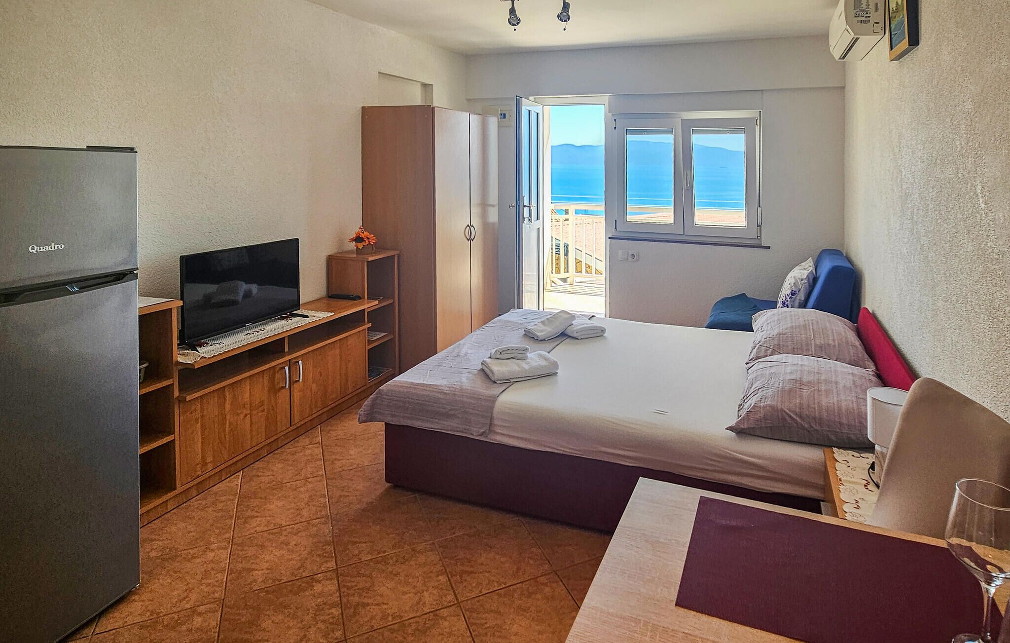 Une chambre d’hôtel de taille réduite, équipée d’un lit, d’une télévision, d’un mini-bar et offrant une vue sur la mer par une fenêtre.