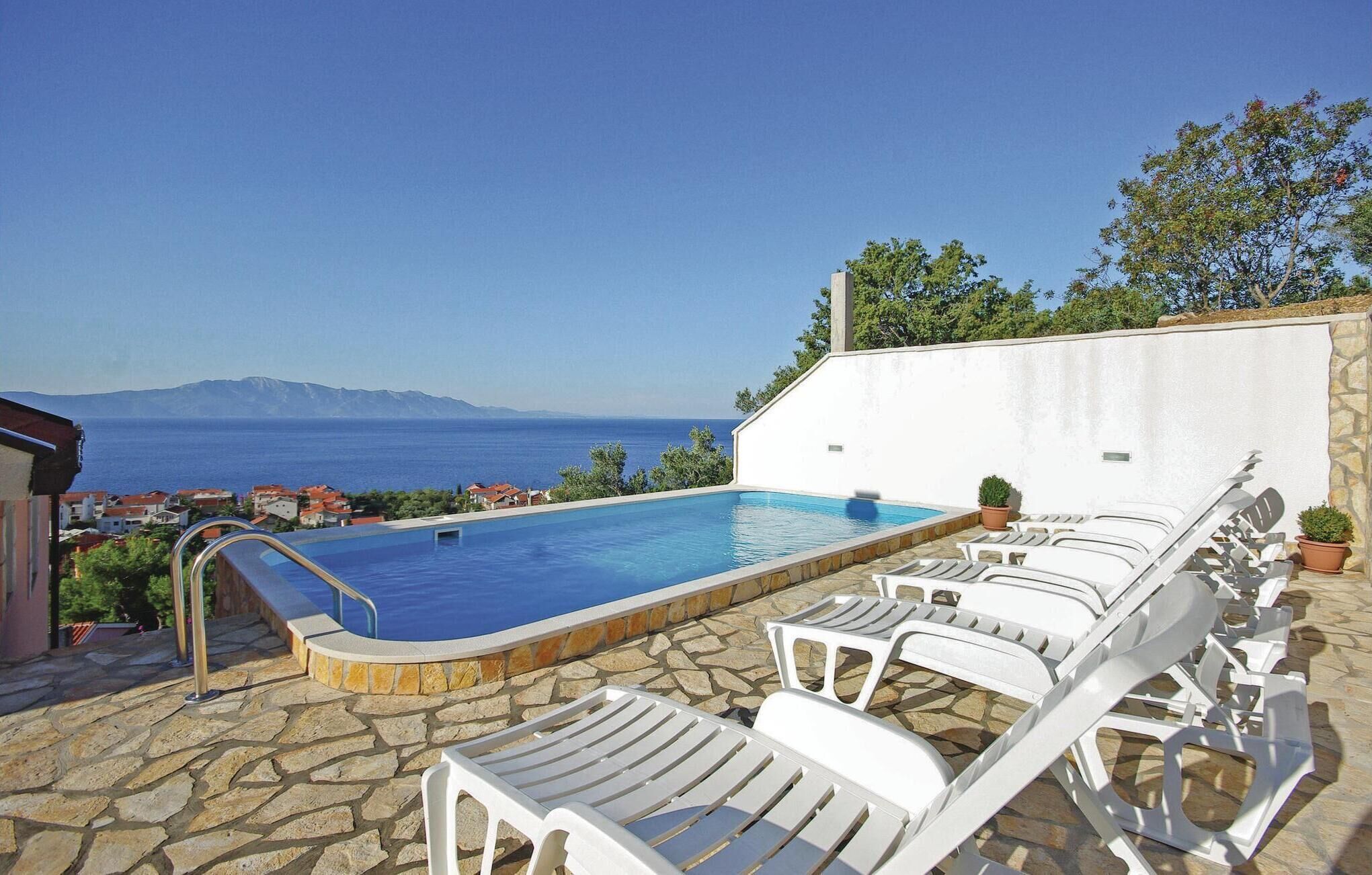 A pool with white lounge chairs, a stone patio, and a view of the sea and mountains.