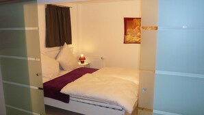 A bedroom with a bed, a nightstand, a lamp, a painting, and a window with curtains. - VV 21; 3-space; mezzanine; Balcony; 78m² - Villa Vogelsang (Sierksdorf)