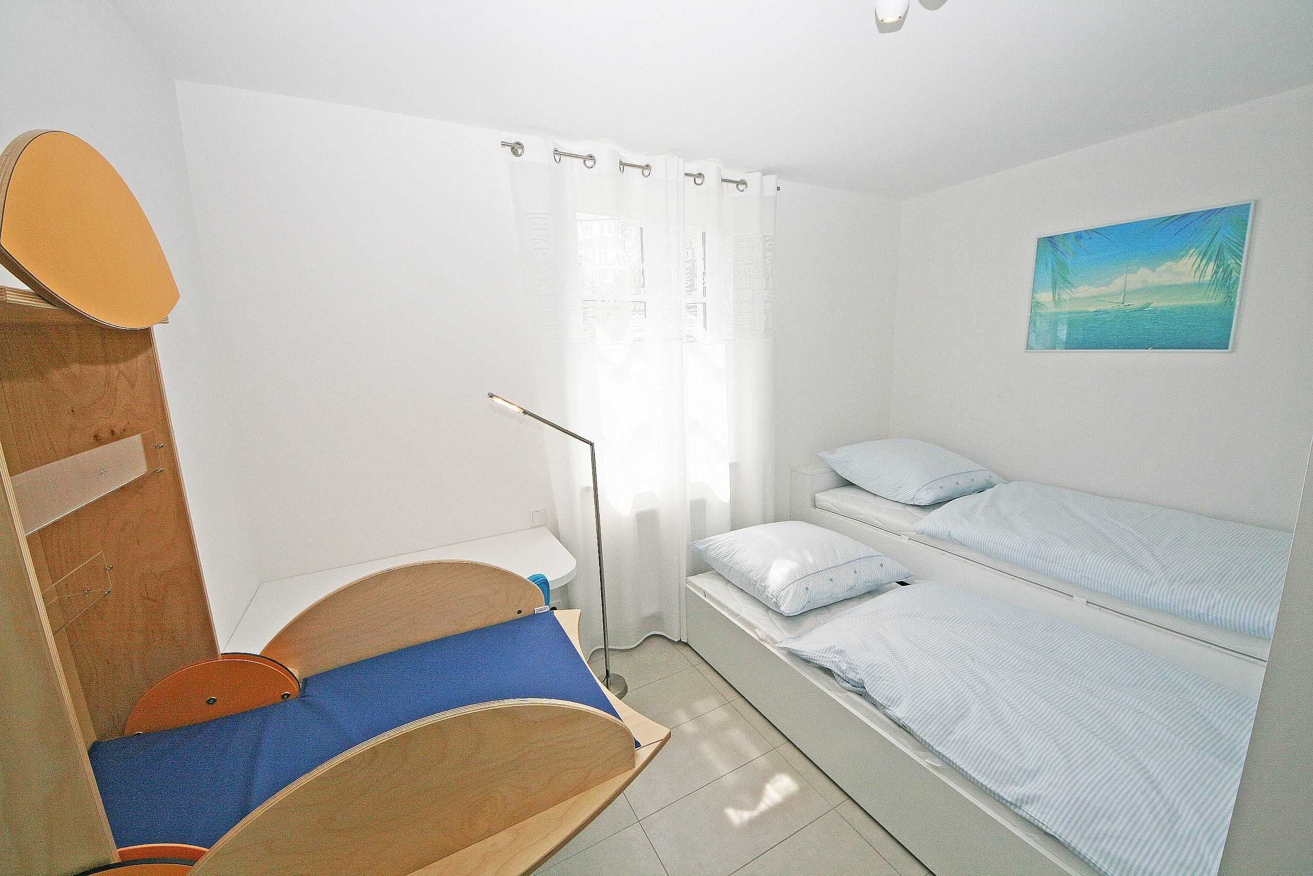 A small, clean room with a single bed, a wooden cabinet, a small table, and a window with curtains.