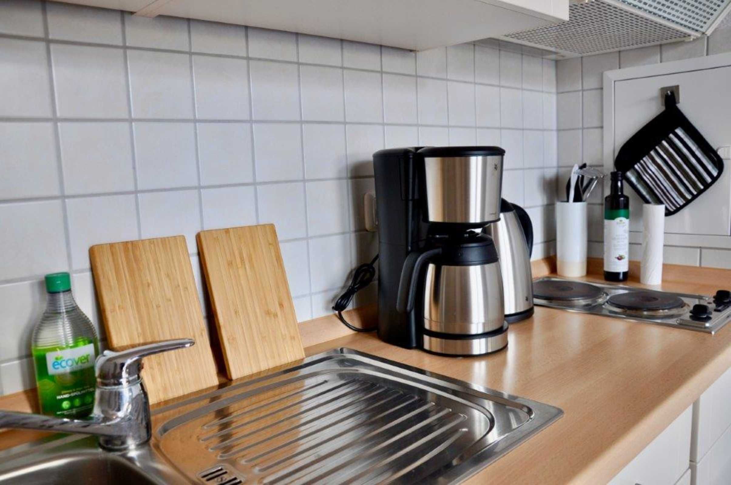 A kitchen with a stainless steel coffee maker, a cutting board, a bottle of dish soap, and a frying pan on the counter.