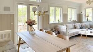 A spacious living room with a wooden dining table, a sofa, and a coffee table. There are multiple windows providing natural light, and a chandelier hangs from the ceiling.