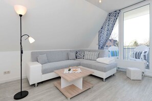 Un salon moderne avec un canapé d’angle, une table basse, un lampadaire et une grande fenêtre donnant sur l’extérieur.