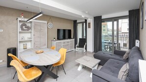Un salon moderne avec un canapé gris, des fauteuils jaunes, une table à manger ronde et un téléviseur posé sur un meuble blanc.
