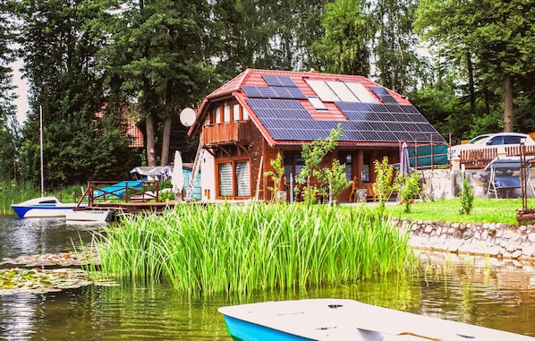Une maison équipée de panneaux solaires, un quai avec des bateaux et un étang bordé d’herbes hautes.