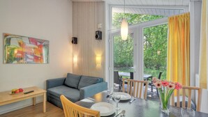 Una sala de estar moderna con un sofá gris, una mesa de comedor de madera para dos personas y un ventanal con cortinas amarillas.