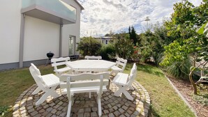 En terrasse med et rundt bord og hvide stole, omkranset af en murstenskant, foran et moderne hus med en glasbalkon.