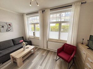 A modern living room with a grey sofa, a wooden coffee table, a red chair, and a large window with white curtains.