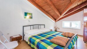 A bedroom with a sloped ceiling, a bed with a colorful checkered bedspread, a white pillow, and a striped pillow. There is a chair, a small bedside table, and a window with a view of trees.