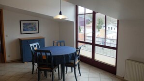 En spiseplads med et rundt bord og stole, et blåt skab, en hængende lampe og et stort vindue med udsigt til bygninger.