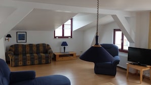 En rummelig loftrum med højt til loftet, en sofa, en lænestol, et fjernsyn og en hængende lampe.
