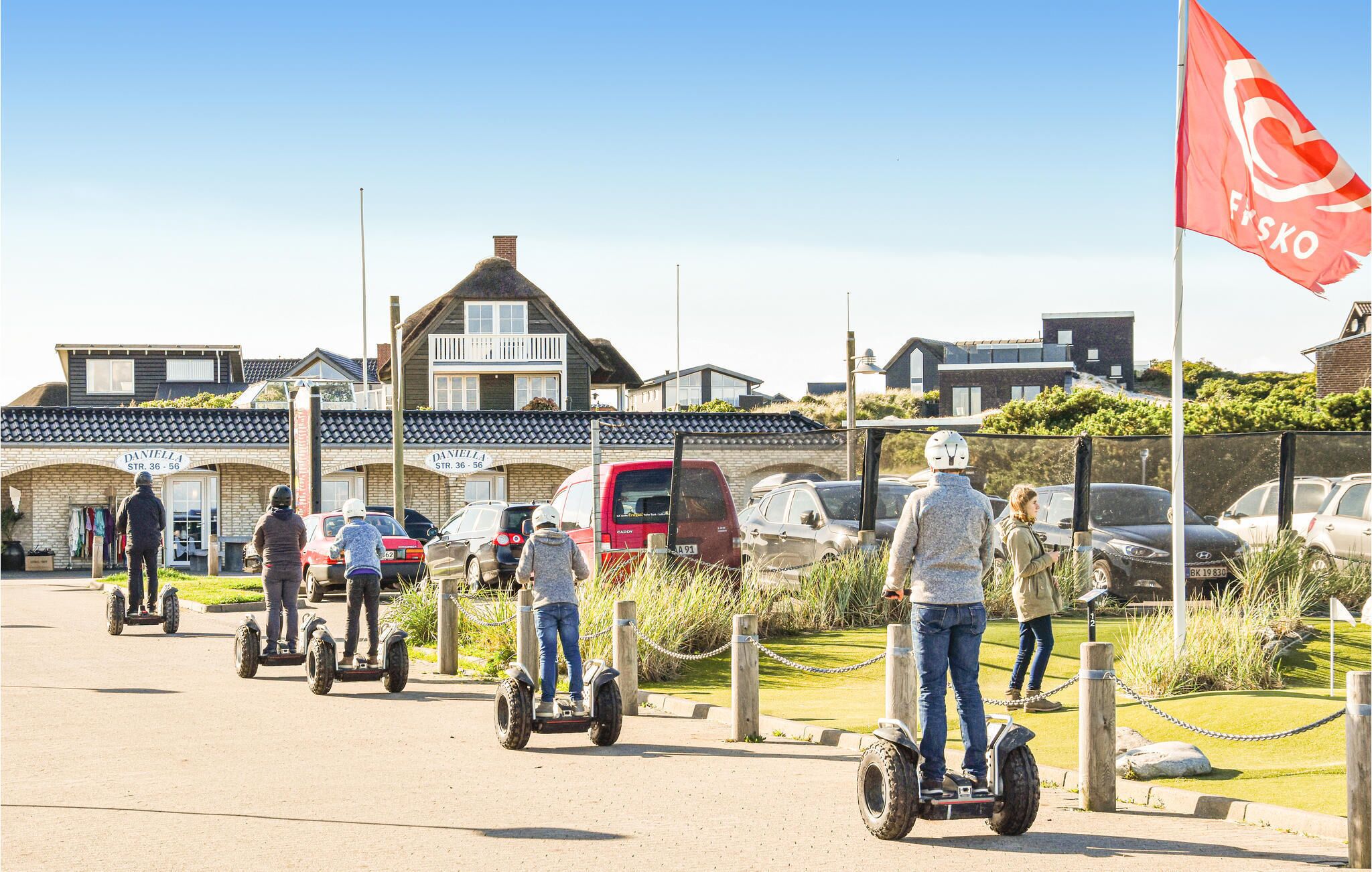 Menschen fahren auf Segways auf einem befestigten Weg, im Hintergrund sind Gebäude und eine Flagge zu sehen.