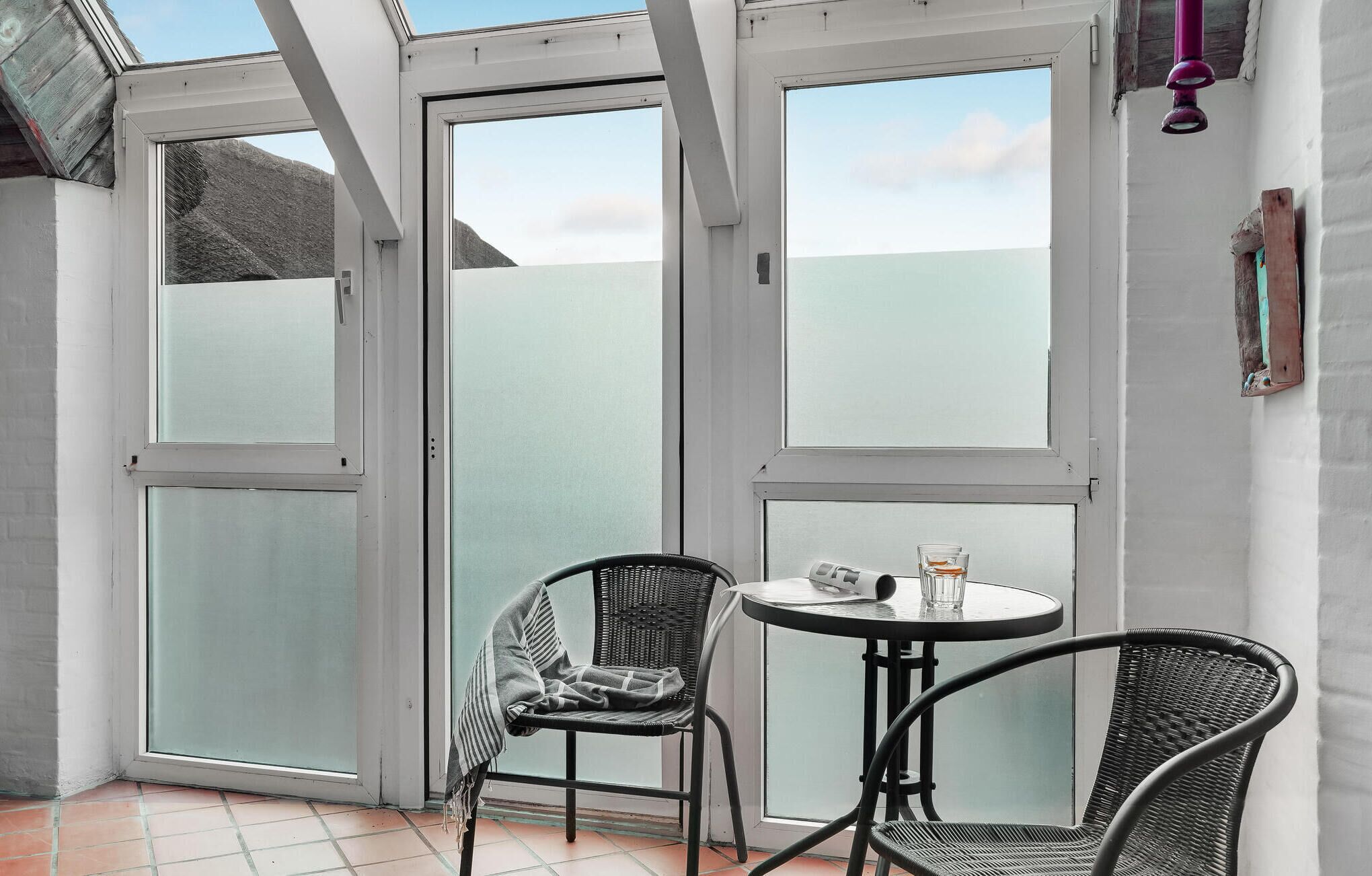 Ein kleiner Patio-Bereich mit zwei Korbstühlen und einem runden Tisch, mit großen Glasüren und Blick zum Himmel.