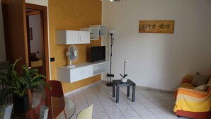 En stue med sofa, fjernsyn, ventilator, lille bord og en potteplante.
