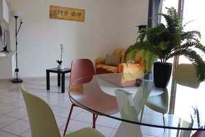 Ein modernes Wohnzimmer mit einem Glastisch, einem roten Sessel, einem Sofa mit orangefarbenen Kissen, einem Topf-Farn und einem Bild an der Wand.
