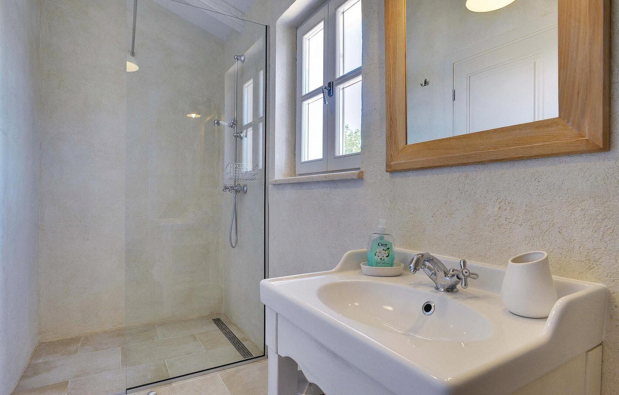 A bathroom with a glass shower enclosure, a white sink with a mirror above it, and a window with a wooden frame.