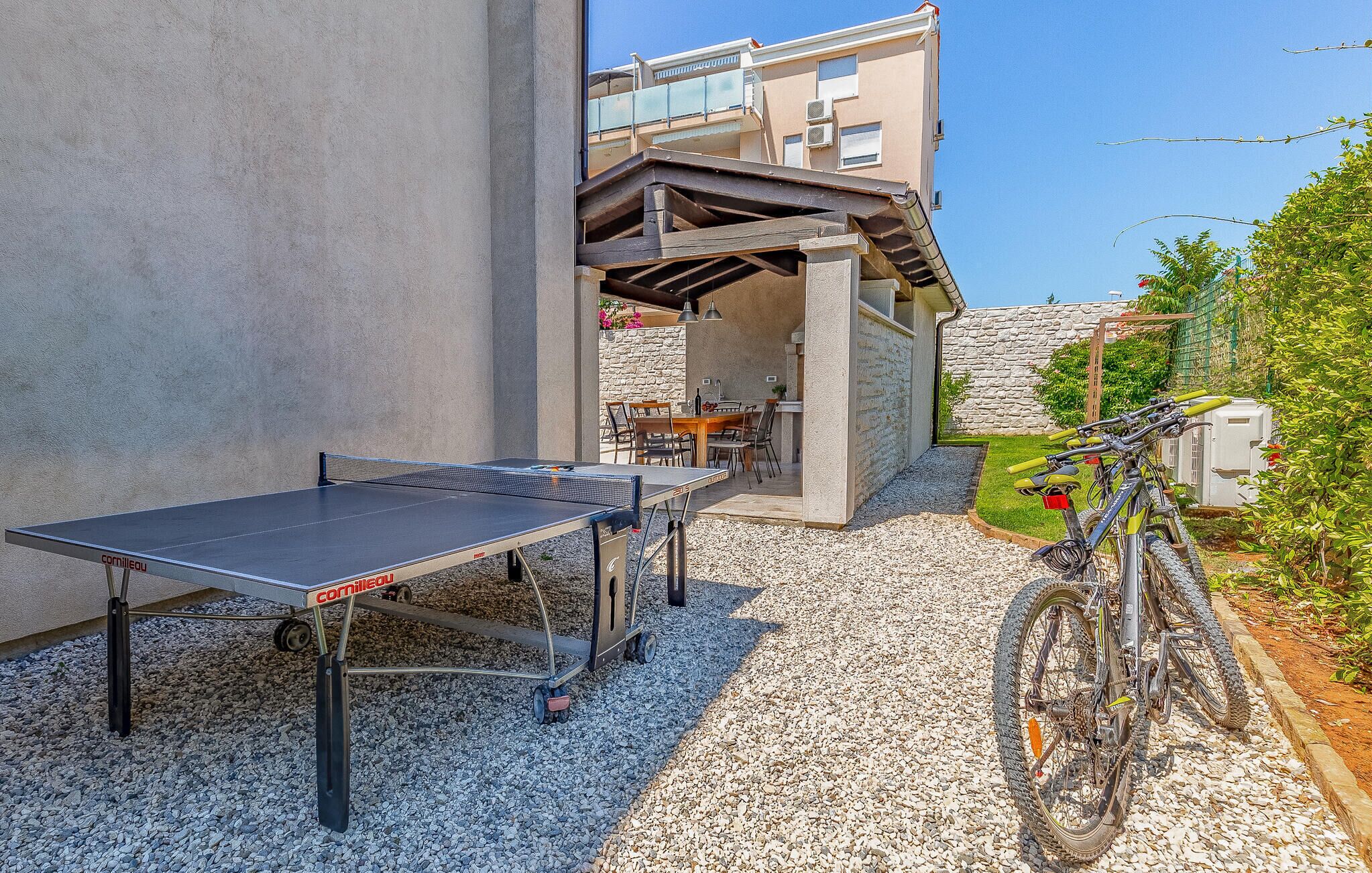 An outdoor table tennis table with a net, a dining table with chairs, and a bicycle parked nearby.