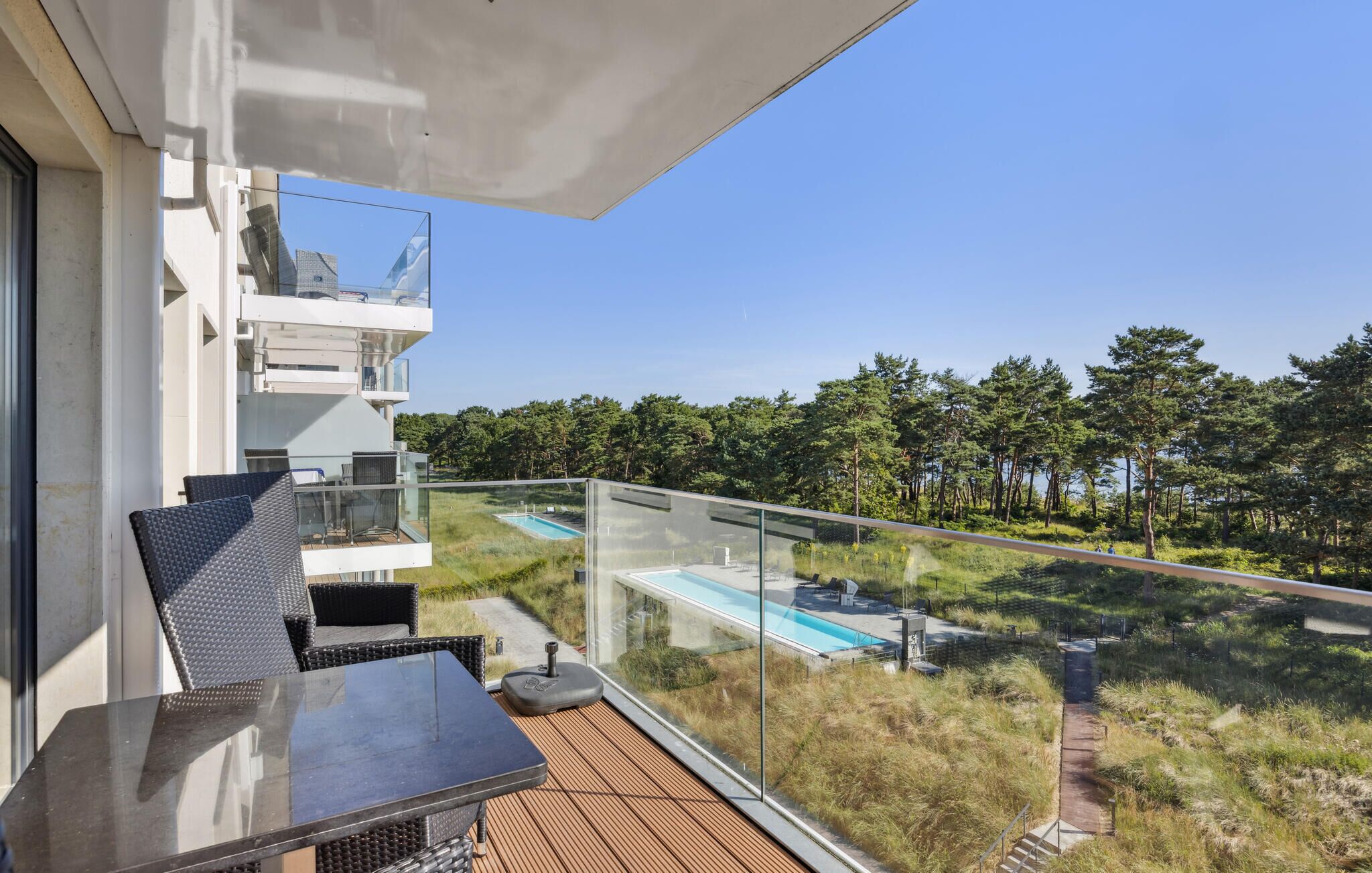 A balcony with a glass railing overlooking a swimming pool and a forested area.