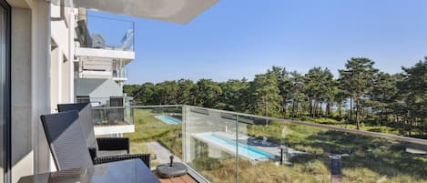 A balcony with a glass railing overlooking a swimming pool and a forested area.