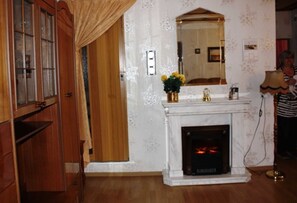 Una habitación con un armario de madera, una chimenea blanca con fuego visible y un espejo sobre la chimenea, además de un estampado de flores en el papel tapiz.