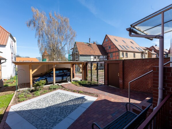 A residential property with a car parked under a covered area, a gated entrance, and a paved patio with a patterned tile design.