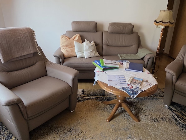 Ein Wohnzimmer mit zwei Sesseln, einem Sofa und einem kleinen Tisch mit Büchern und Papieren.