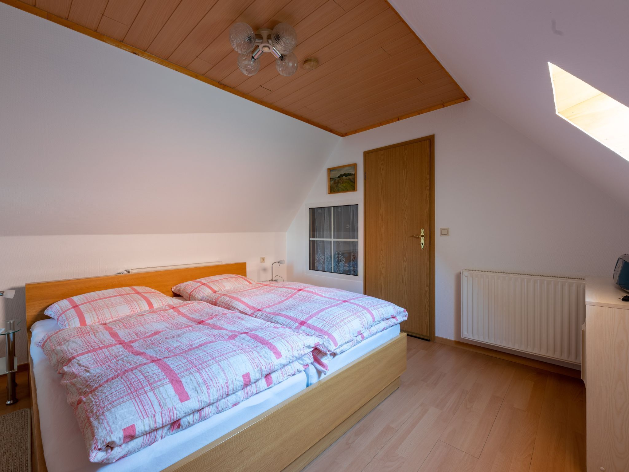 Een slaapkamer met een houten plafond, een bed met een beddengoed met een patroon, een raam met gordijnen, een radiator en een houten garderobe.