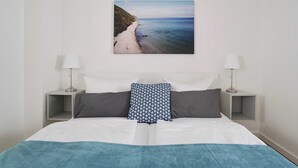 Un lit bien fait, recouvert d’une couvre-lit blanche et bleue, avec deux oreillers gris et un coussin décoratif à motifs. On y trouve deux tables de chevet avec des lampes, et un tableau encadré représentant une scène de plage, accroché au mur.
