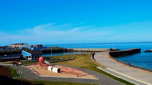 Un paseo marítimo con un parque infantil, un edificio azul y un muelle que se adentra en el agua.