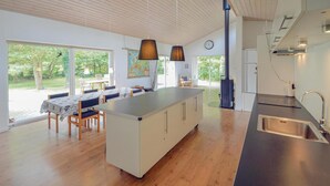 Une cuisine moderne dotée d’un îlot central, d’un coin repas avec une table et des chaises, et de grandes fenêtres offrant une vue sur l’extérieur.