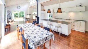 Une cuisine moderne avec des meubles blancs, un îlot central, des luminaires suspendus et un coin repas agrémenté d’une nappe à motifs.