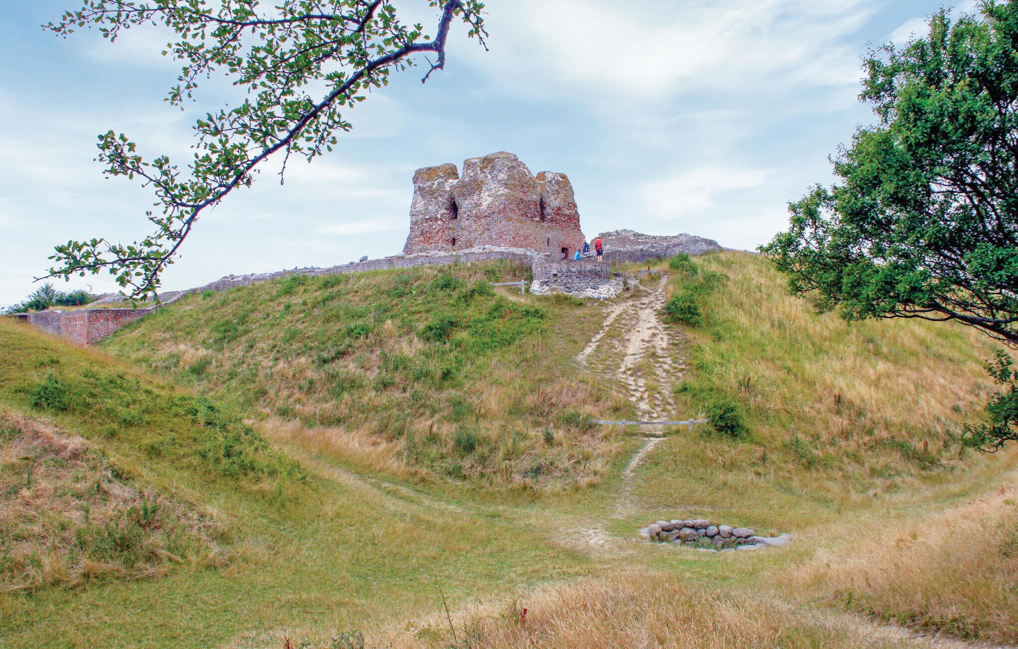 Eine historische Steinarchitektur auf einer grasbewachsenen Hügelkuppe unter blauem Himmel.