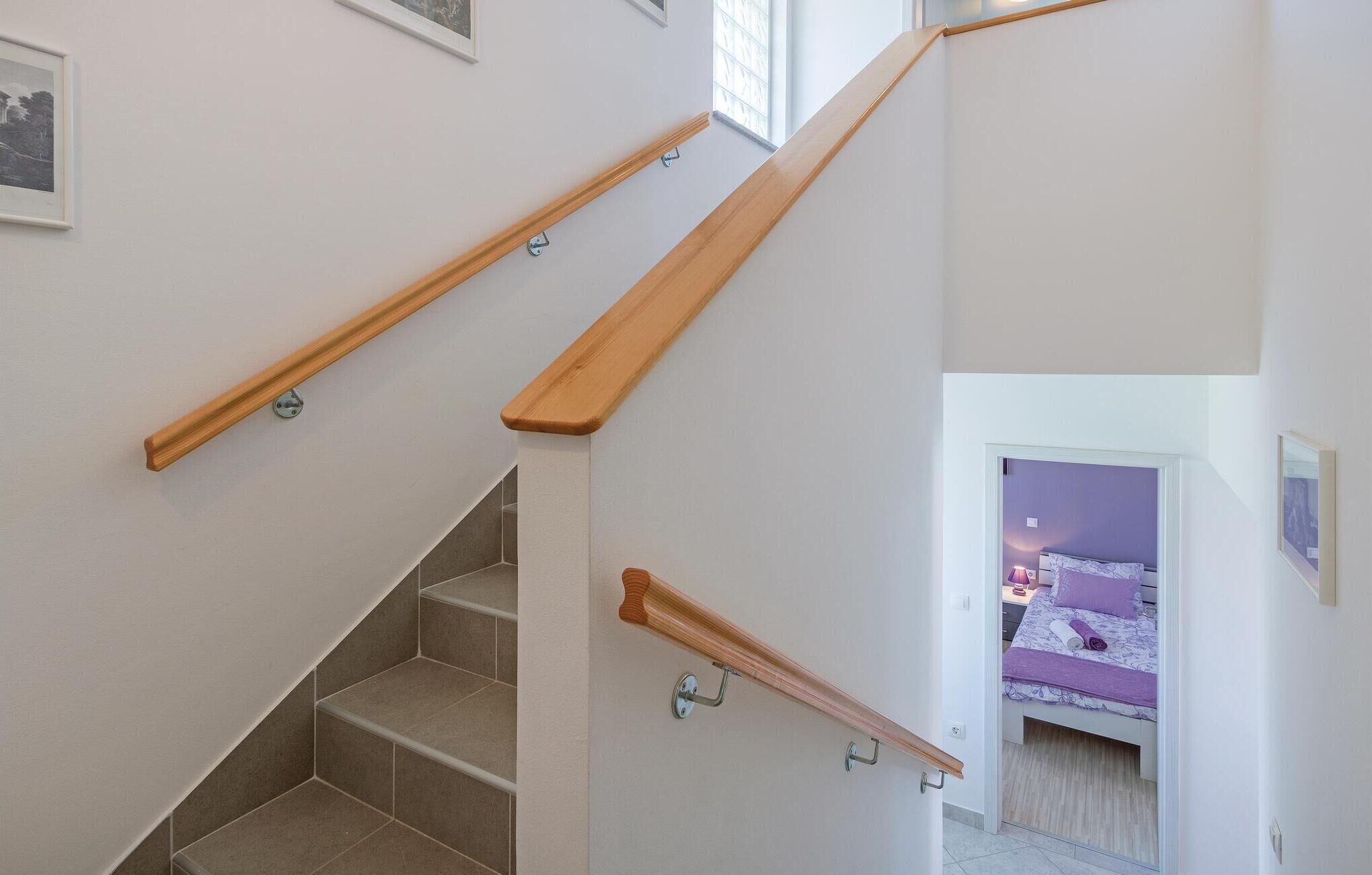 Una escalera con barandillas de madera, un dormitorio con cama y ropa de cama morada, y una ventana que deja entrar luz natural.