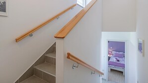 Una escalera con barandillas de madera, un dormitorio con cama y ropa de cama morada, y una ventana que deja entrar luz natural.