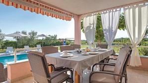 Un área de comedor exterior cubierta con vista a la piscina, sillas de mimbre y una mesa dispuesta para una comida.